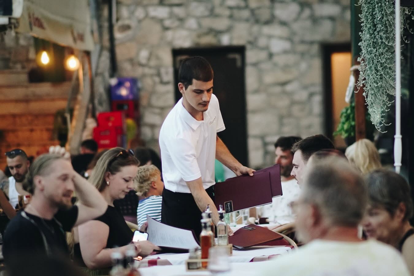 Konobar u bijeloj polo majici drži tamni jelovnik pokraj gostiju za stolovima na terasi uz kamenu fasadu